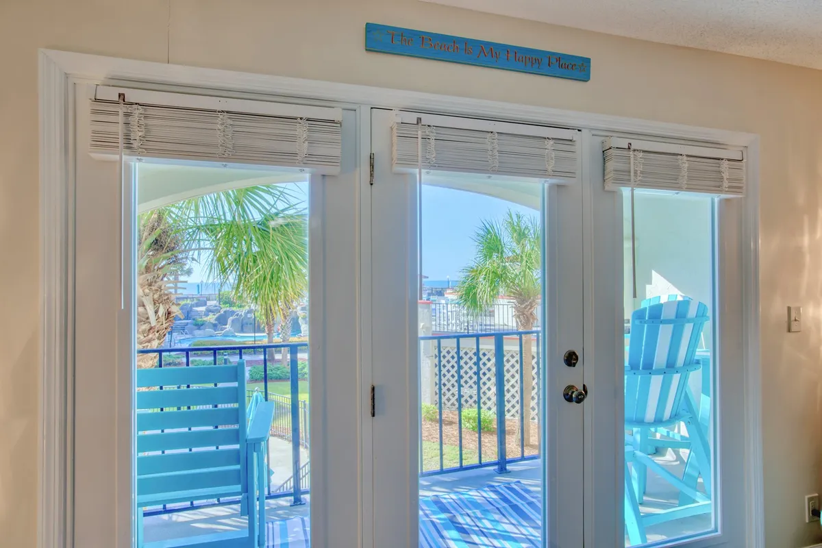 A bright interior view with double doors open to the balcony, two blue chairs positioned for conversation or enjoying the view, and palm trees visible just beyond. Sunlight streams through, filling the space with a welcoming glow.