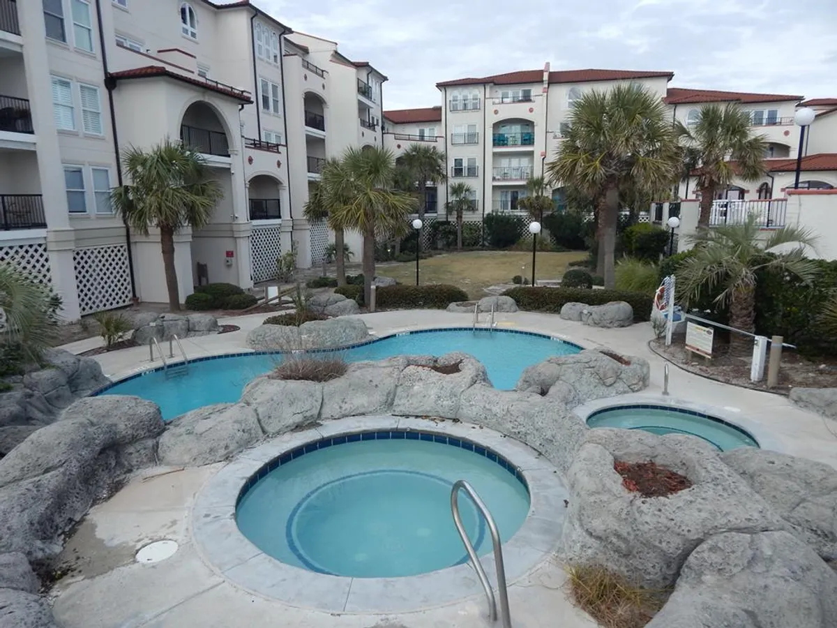 A sunlit shot of the resort’s hot tubs framed by natural rock formations and swaying palm trees, with glimpses of surrounding Mediterranean-style buildings. Guests are seen relaxing in the steaming tubs, reinforcing the theme of indulgence and relaxation.