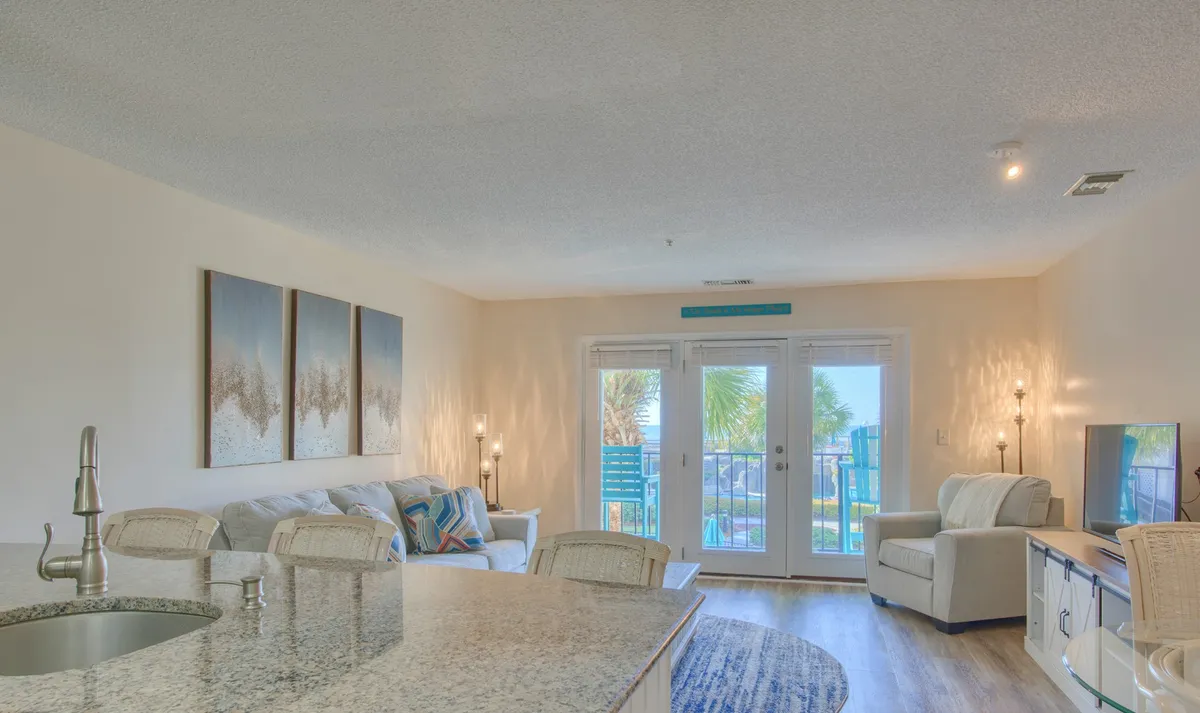 A welcoming open living space with a granite countertop separating the kitchen from the seating area, large windows that bathe the room in sunlight, modern artwork on the wall, and a television positioned for easy viewing. The balcony is visible, suggesting outdoor living is just steps away.