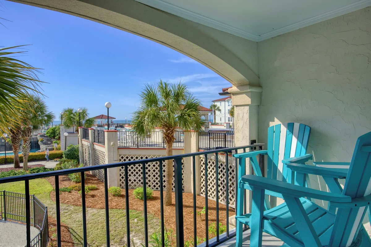 The image features a tranquil condo balcony outfitted with inviting chairs, framed by swaying palm trees. Guests can look out over the resort's sparkling pool with the vast Atlantic Ocean as the backdrop, all bathed in gentle natural light. The scene embodies leisure and the feeling of being transported to a beachside retreat.
