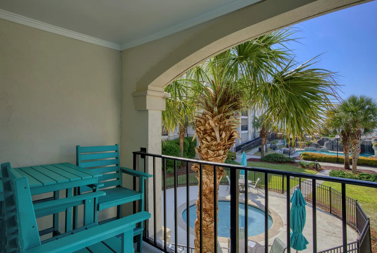 This exterior image features inviting teal outdoor chairs overlooking a sparkling swimming pool, with lush palm trees framing the scene. The tranquil setting is enhanced by the soft evening light, hinting at both sunrise and sunset views from the balcony. The overall mood is peaceful and perfect for quiet solo retreats or unwinding after a day of local exploration.