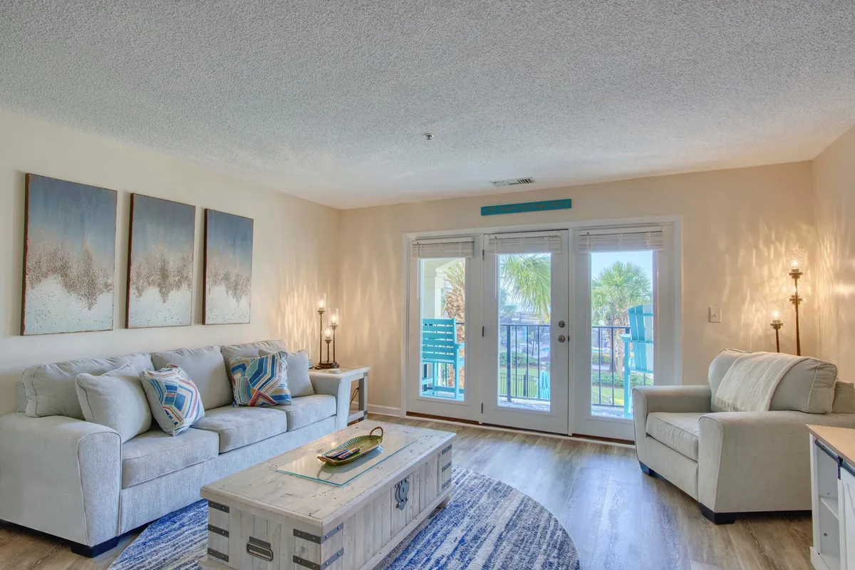 A bright, cheerful living room featuring a plush sofa with colorful cushions, a modern coffee table adorned with decorative items, and sliding glass doors that open directly to a private balcony. Natural light floods the space, enhancing the inviting atmosphere and drawing attention to the seamless indoor-outdoor flow.