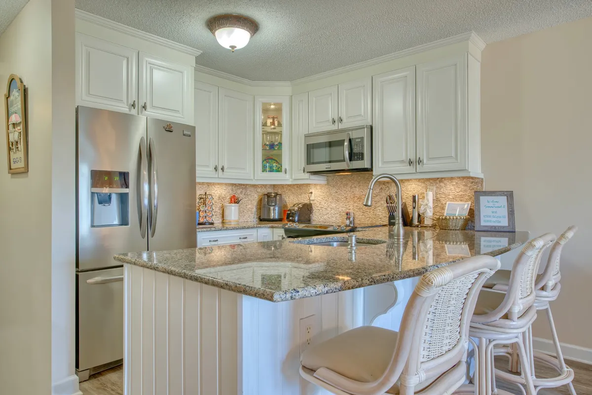 A welcoming kitchen space featuring gleaming granite countertops, modern stainless steel appliances, crisp white cabinetry, and comfortable bar stools at the breakfast bar. The layout is open and inviting, perfect for meal prep or casual dining.