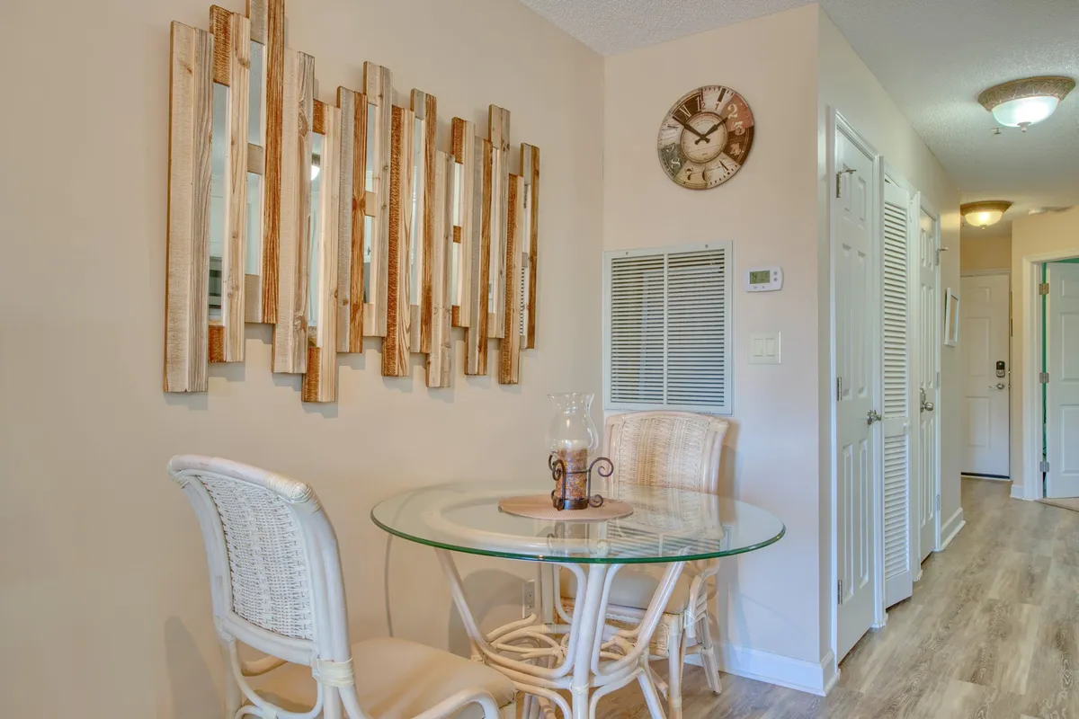 A cozy dining area featuring a round glass table surrounded by wicker chairs, set near a wooden mirror and wall clock. Soft morning light streams in, creating an intimate spot for shared meals or morning coffee.