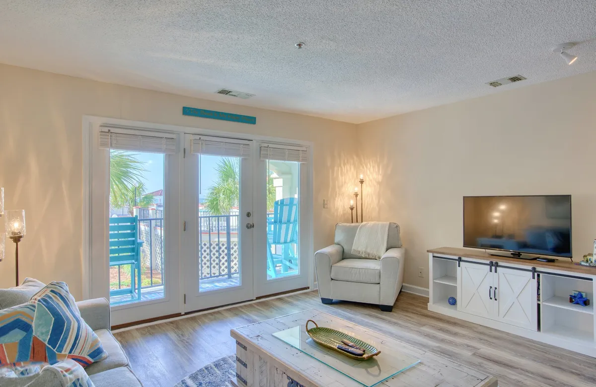 A relaxed seating area with a comfortable armchair, a flat screen TV, a chic coffee table with stylish decor, and sliding glass doors that lead out to the balcony. Subtle morning light streams through, enhancing the tranquil, inviting mood of the living space.