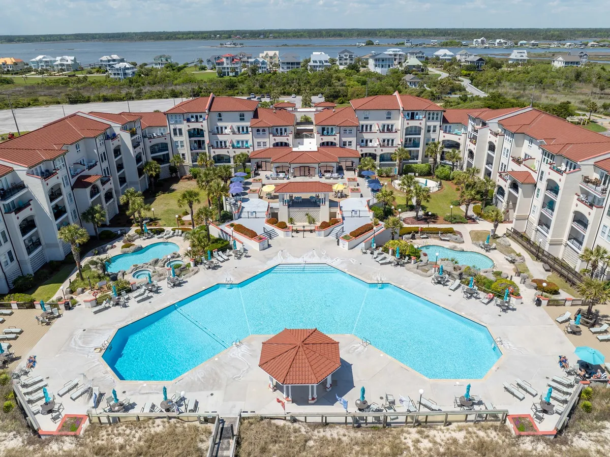 This image captures a large swimming pool surrounded by palm trees, colorful umbrellas, and the Villa Capriani complex buildings in the background. The inviting pool area is lively and sunlit, with a relaxed, cheerful atmosphere that hints at vacation leisure and social gatherings.