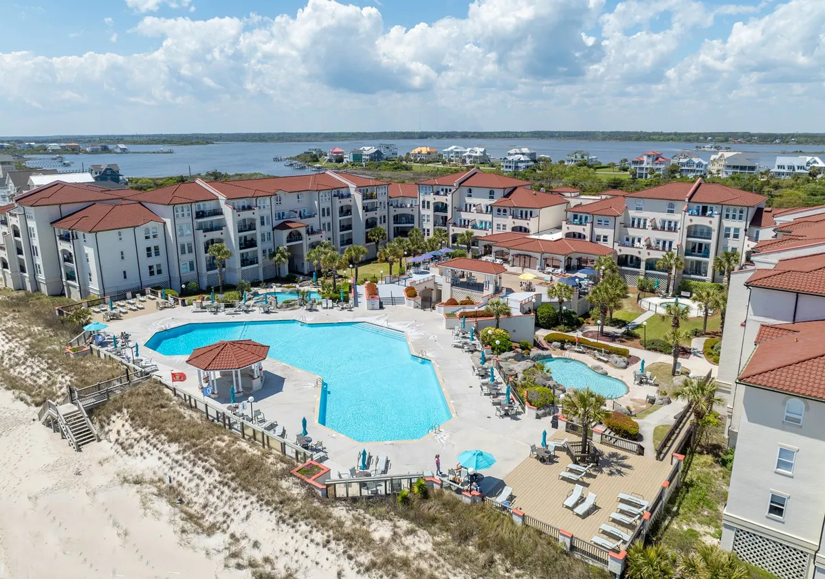 A bright, cheerful view of a large swimming pool bordered by palm trees and colorful umbrellas. The deck area features ample lounge chairs, and families are seen gathering for poolside fun, while the nearby beach can be glimpsed in the distance.