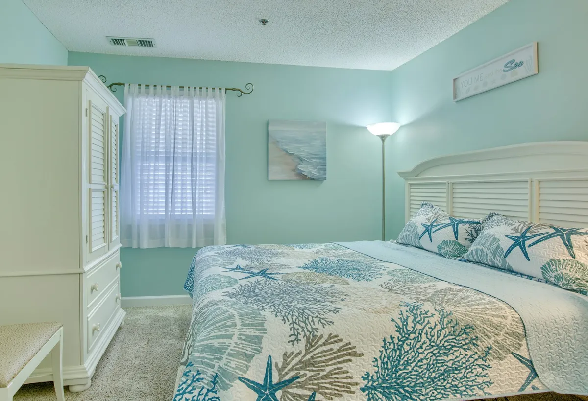 A peaceful bedroom with a king-sized bed dressed in coral-patterned bedding, accompanied by crisp white furniture and calming artwork on the wall. The space feels light-filled and serene, designed to encourage restful sleep and relaxation.