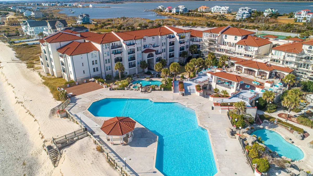 This exterior image captures a sweeping view of the Villa Capriani’s resort-style pool complex. The frame is filled with large swimming pools, lush palm trees, colorful resort buildings, and a direct glimpse of the beachfront, all teeming with activity. Poolside lounge areas and joyful families emphasize the inviting, vacation atmosphere.