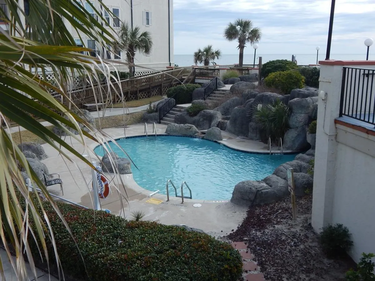 A sparkling swimming pool framed by natural rock formations, waving palm trees, and a neat walkway, creating an inviting oasis atmosphere ideal for family relaxation and fun. The ample pool area is perfect for both energetic play and calm lounging, reflecting the resort's appeal to families with children of all ages.